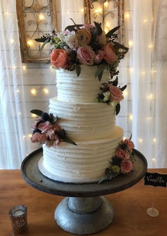 Three-tiered white cake decorated with flowers on a wooden table with string lights in the background.